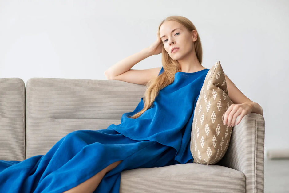 A woman wearing a flowing royal blue dress with a high slit sits on a beige sofa, resting her head against one hand and leaning slightly into an ornate patterned pillow.