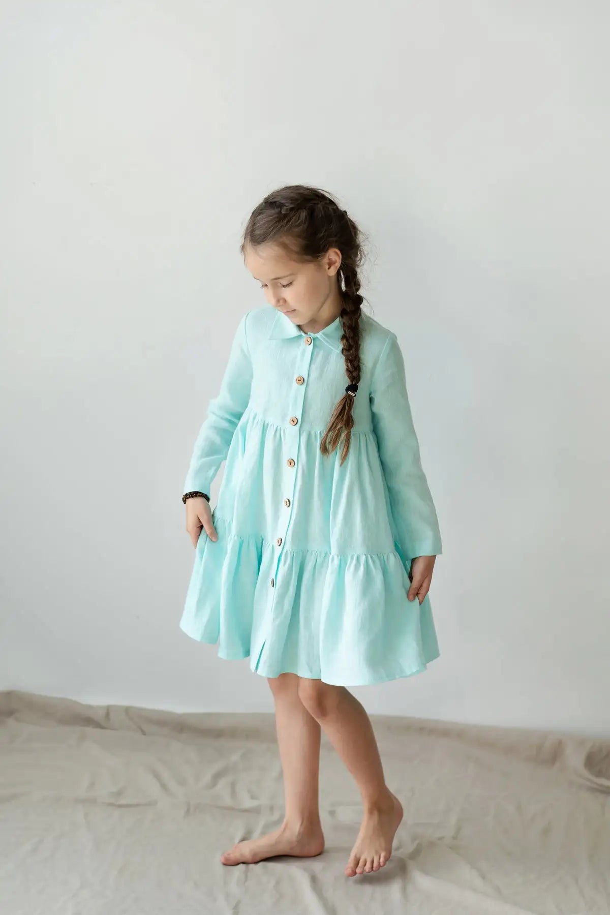 A young girl wears a light mint green, long-sleeved tiered dress with a button-front and collar.